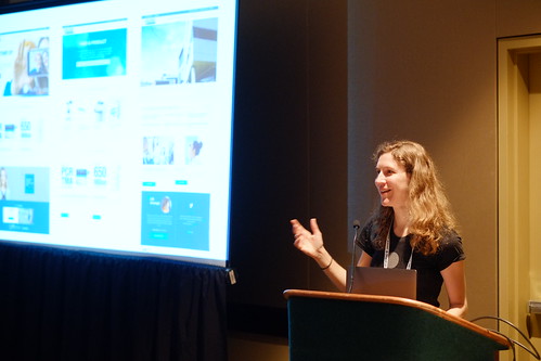 Photo of a white woman presenting from a podium, there is a screen with a slide projection in the background.